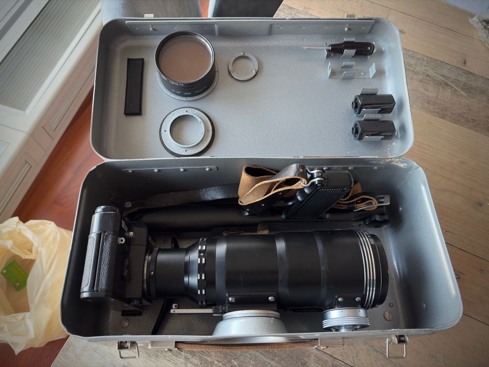 A camera with a shoulder stock and pistol grip mounted to a large lens and its metal carrying case