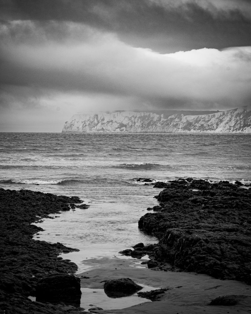 A rocky shore with white chalk cliffs across the water under whisps of low cloud