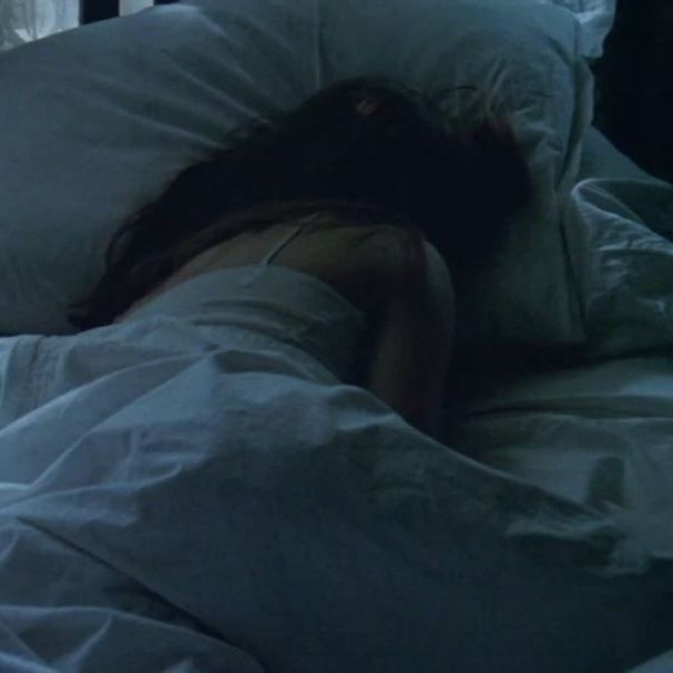 Una chica hecha una bolita en la cama, tapada con el edredón blanco y la cara hundida en la almohada.