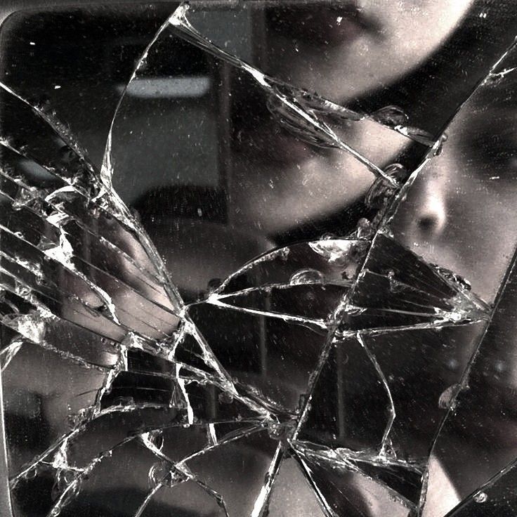 Una foto de una chica reflejada en un espejo roto. La chica apenas se ve, y la imagen en sí es muy oscura.