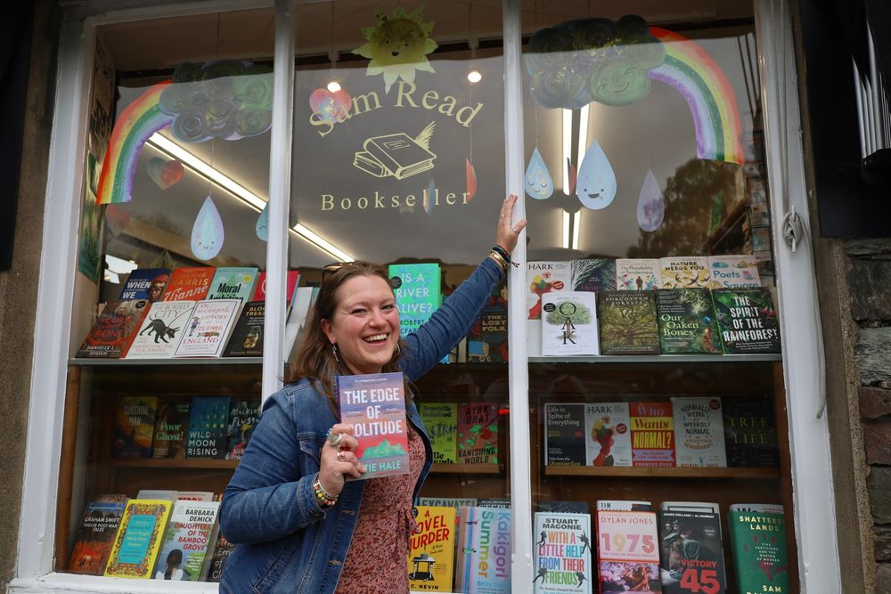 Katie, a white woman with long brown hair wearing a denim jacket, leans on the window of Sam Read, smiling, and holding a paperback book 'the edge of solitude'. The window is full of colourful new hardbacks, with rainbows, raindrops and a smiling sun hanging above them. 