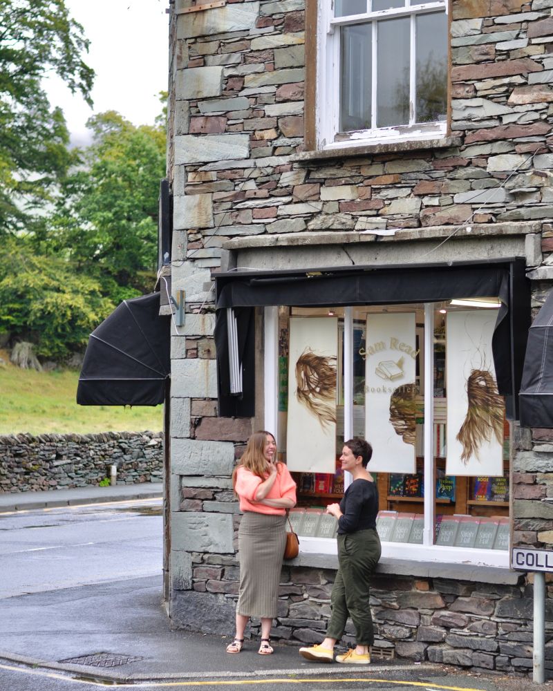 bookshop corner with Sarah and Jocelyn laughing and talking in front of the window with Jocelyn's art and many copies of Helm in, with drystone wall, trees, field and the wet road behind them.