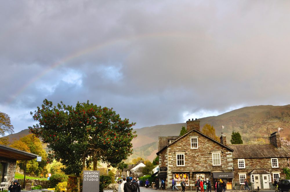 rainbow over broadgate grasmere, with lots of people walking around outside Heaton Cooper, Sam Read Bookseller and Lucia's takeaway, and hazy sunlit fells, with autumn colours on trees. 1st bow is quite strong, 2nd is very faint. 
