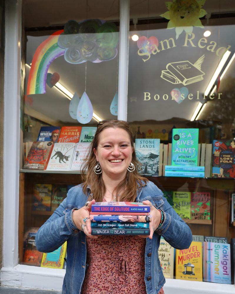Katie smiling and holding book stack outside bookshop window with the Edge of solitude by Katie hale, at the mountains of madness by HP Lovecraft, Frankenstein by Mary Shelley, lean fall stand by Jon McGregor, and The wager and the bear by John ironmonger. 