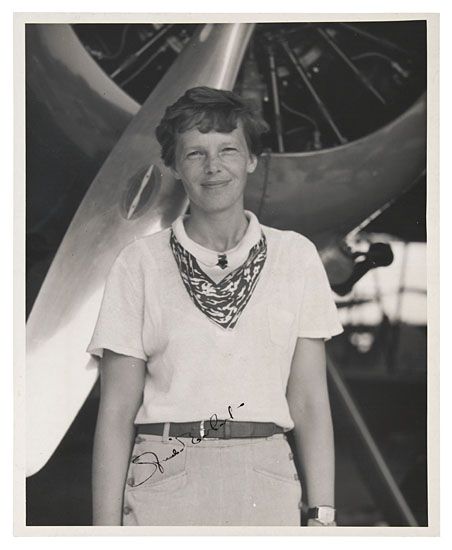 Amelia Earhart standing in front of plane engine while smiling.