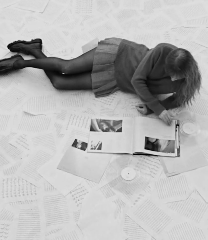 A woman (Taylor Swift) laying on the ground surrounded by paper files and clear vinyls looking at a photo book. The photo is in black & white