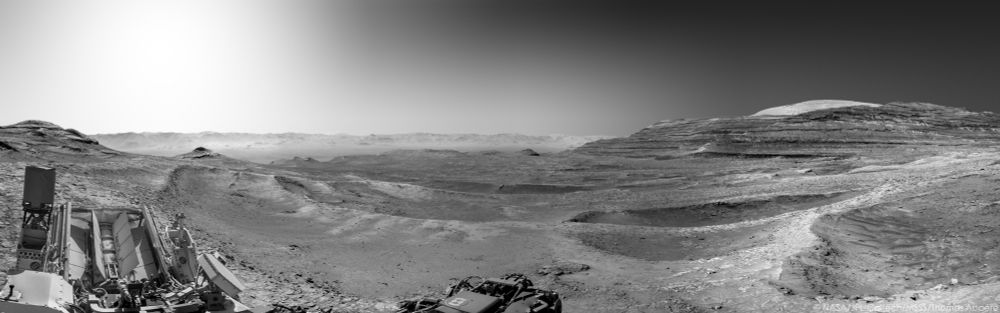 Un paysage martien en noir et blanc. Le rover Curiosity est visible en bas à gauche avec son RTG et ses antennes.