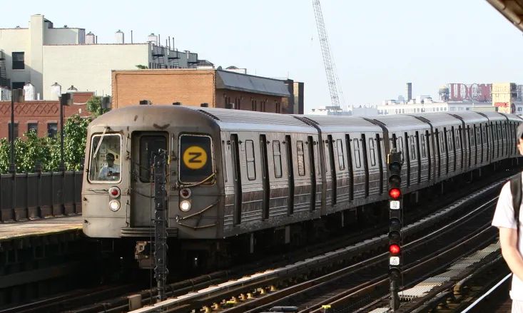 an image of NYC's N train, riding down the rails above ground off to do train things. the normally circular yellow N sign in the window has been edited and turned sideways, to make it look like the letter Z.