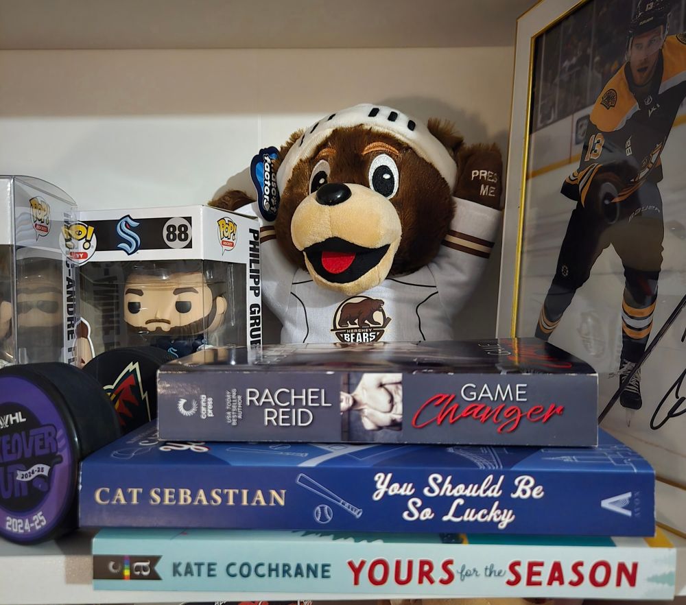 In a horizonally laid stack three books sit from the bottom up:
Yours for the season by kate Cochran
You should be so lucky by cat sebastian
Game changer by rachel reid 

Surrounding the books is various hockey paraphernalia including a hershey bears mascot plush and a couple pucks