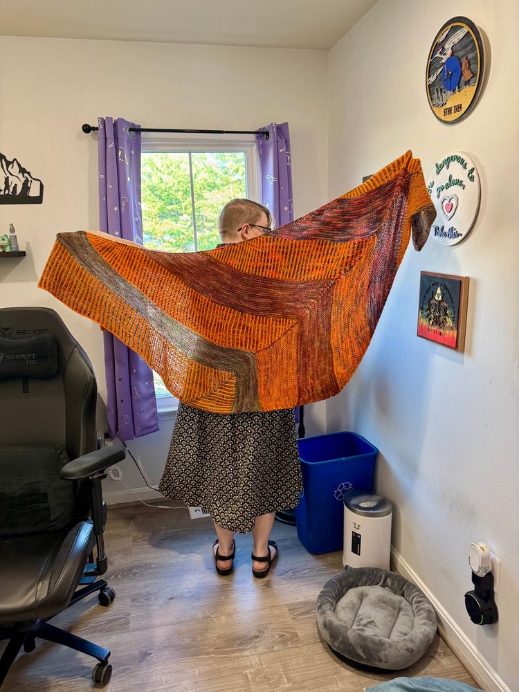 A woman holding a handknit brioche shawl sprawled across her back in oranges, browns, and reds. 