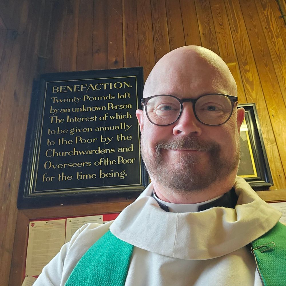 A vicar with an old sign on the vestry wall behind him. This reads "Benefaction. Twenty pounds left by an unknown person  the interest of which to be given annually to the poor by the churchwardens and overseers of the poor for the time being.