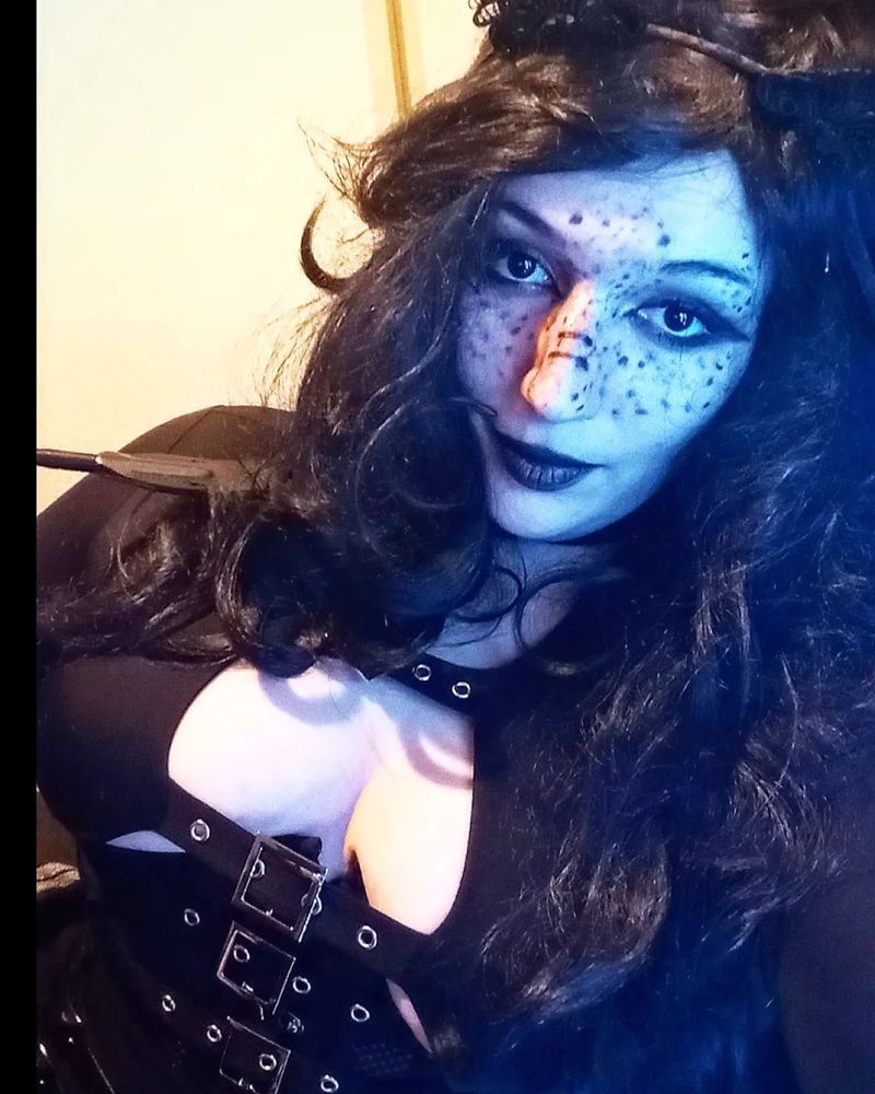 Alliesha Bay is wearing a black wig with a black shirt featuring the cleavage area with an underbust corset. They have Gothic makeup on and black cat ears.