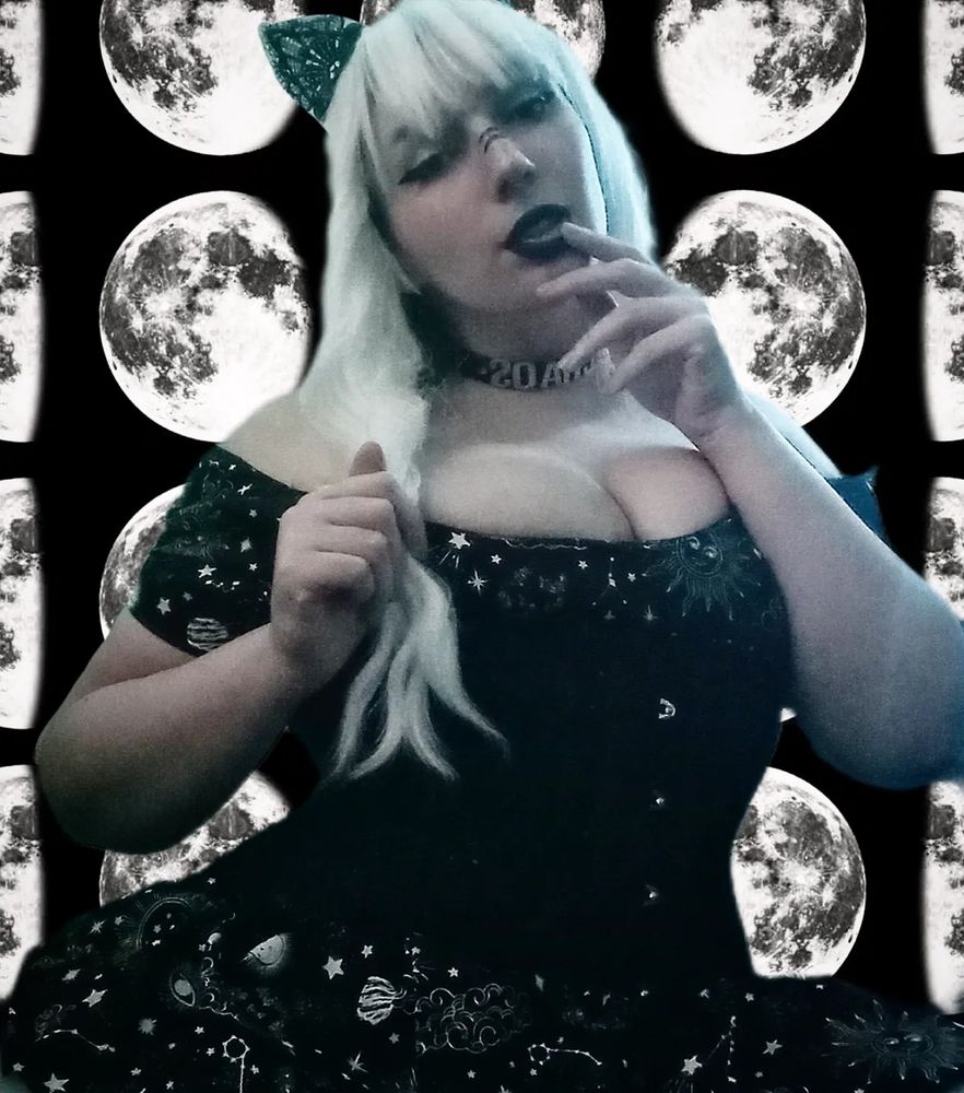 Alliesha Bay is wearing a black and white dress with a black corset and a white wig paired with black lace cat ears and goth makeup. They have a 'chaos' choker and the background is moons. She looks down sensually at the camera while holding her arms up near their face. 