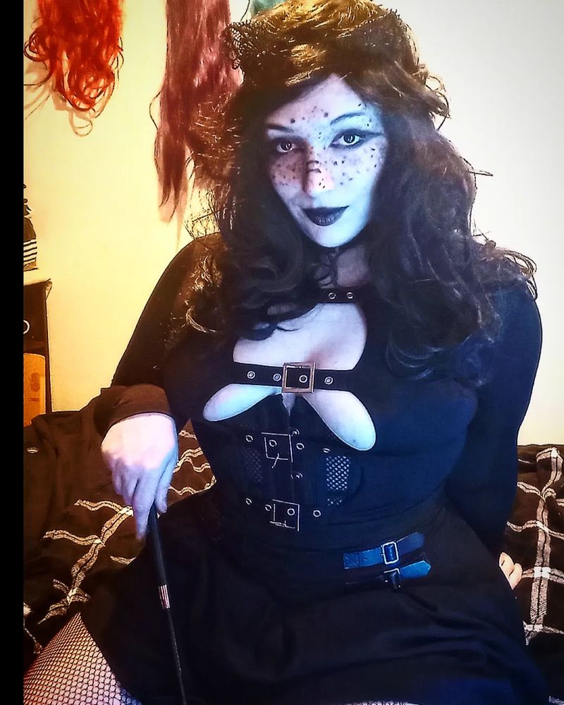 Alliesha Bay is wearing a black mini skirt with a black corset, black long sleeve shirt with cutouts, a long wavy black wig and gothic makeup. They hold a flogger in one hand against their thighs and look seductively at the camera. 