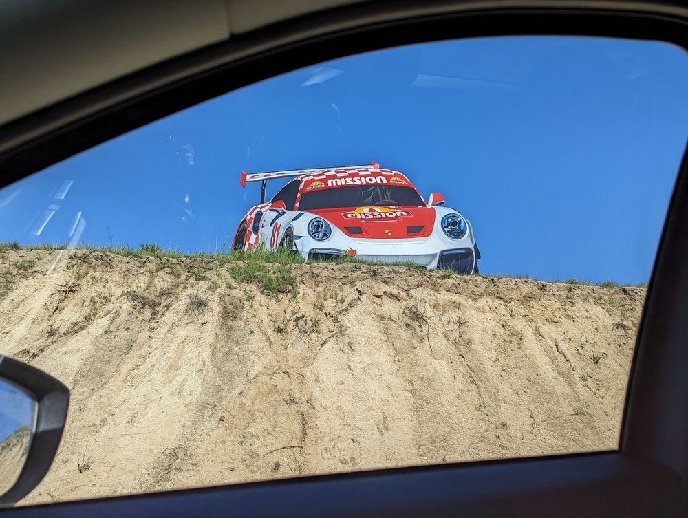 A photo of a billboard version of a Porsche 911 Cup car that has the bottom portion of the billboard partly obscured by a sandy hill. The photo was taken from the inside of a car