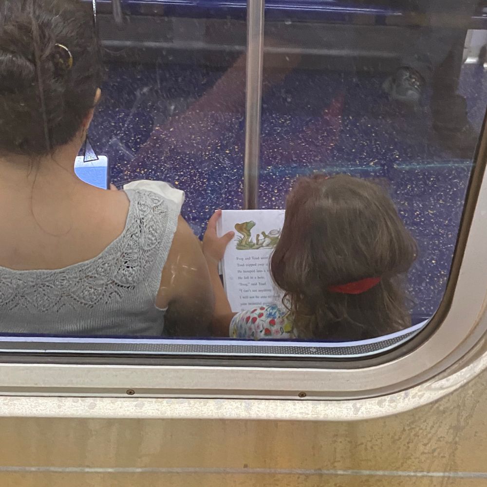 Through the subway window, an over-the-shoulder glimpse of Frog and Toad book in hands of dark-haired girl, sitting beside a dark-haired woman. 