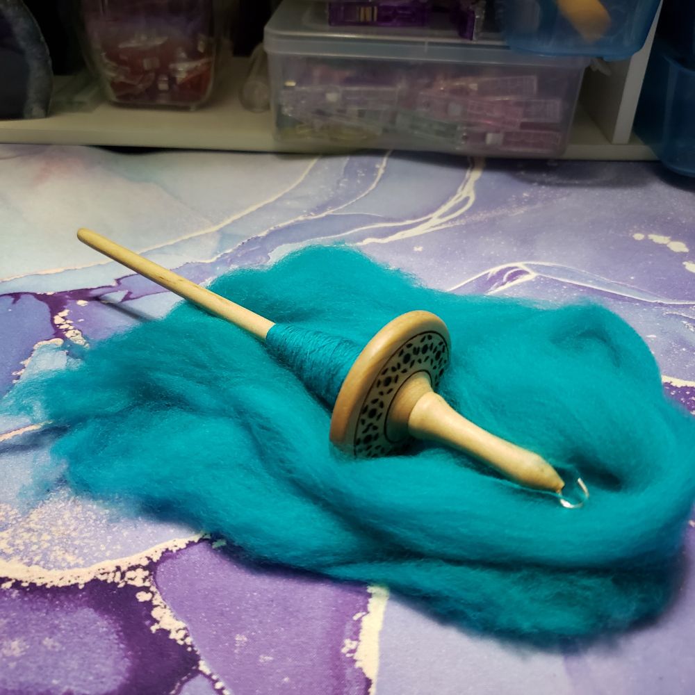 A hand turned wooden drop spindle sits in a small pile of turquoise colored merino wool top on Kelli's desk. The spindle has decorative  pyrographed motifs on the top of the whorl. A small cop of handspun yarn is wound onto the spindle. 