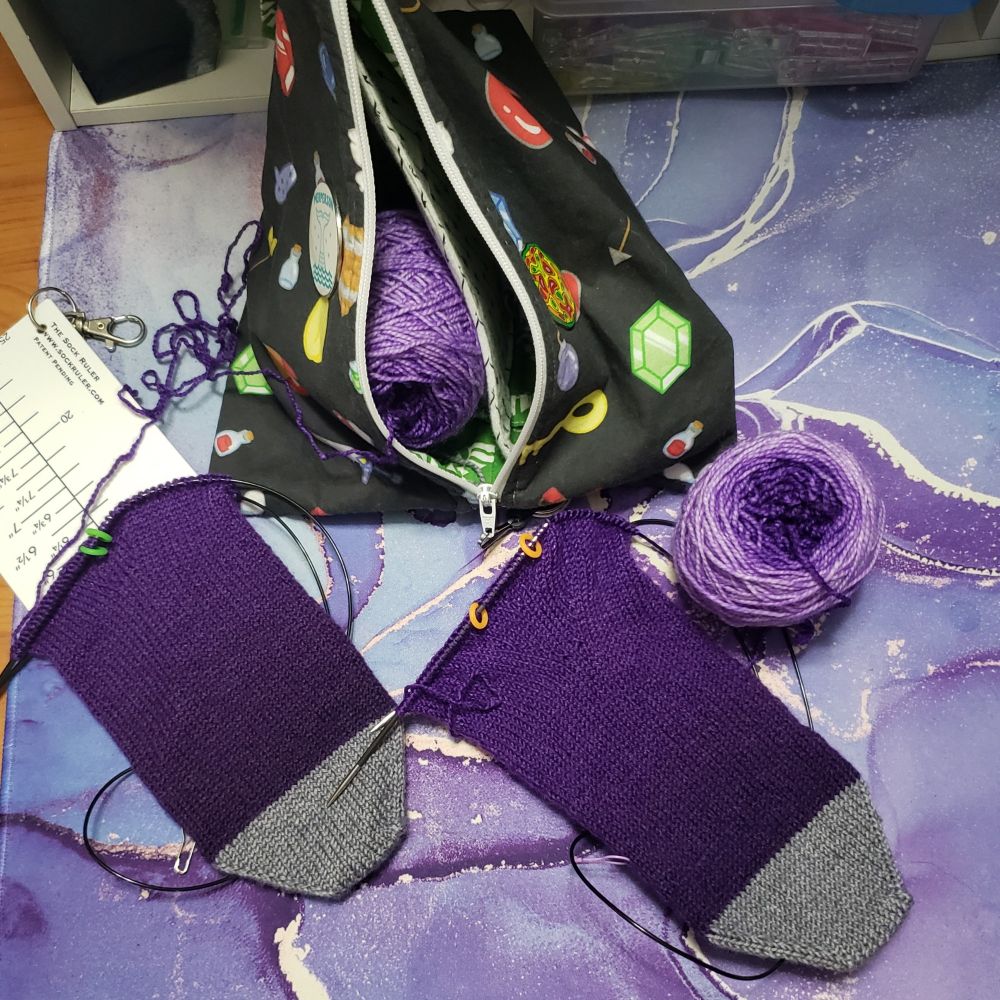 An overhead view of Kelli's desk on which sits a sock knitting project in progress with a zelda themed divided tetrahedral fabric project pouch. The socks are vanilla double gusset on the bottom toe up socks. Both socks are being knit concurrently on separate circular needles. One sock has been placed on a sock ruler. Both socks have semisolid hand dyed medium dark silver grey toes. The foot of the sock is a dark to light semisolid hand dyed purple gradient that was started on the dark end of the gradient. The small center pull balls they are being knit from are visible. 