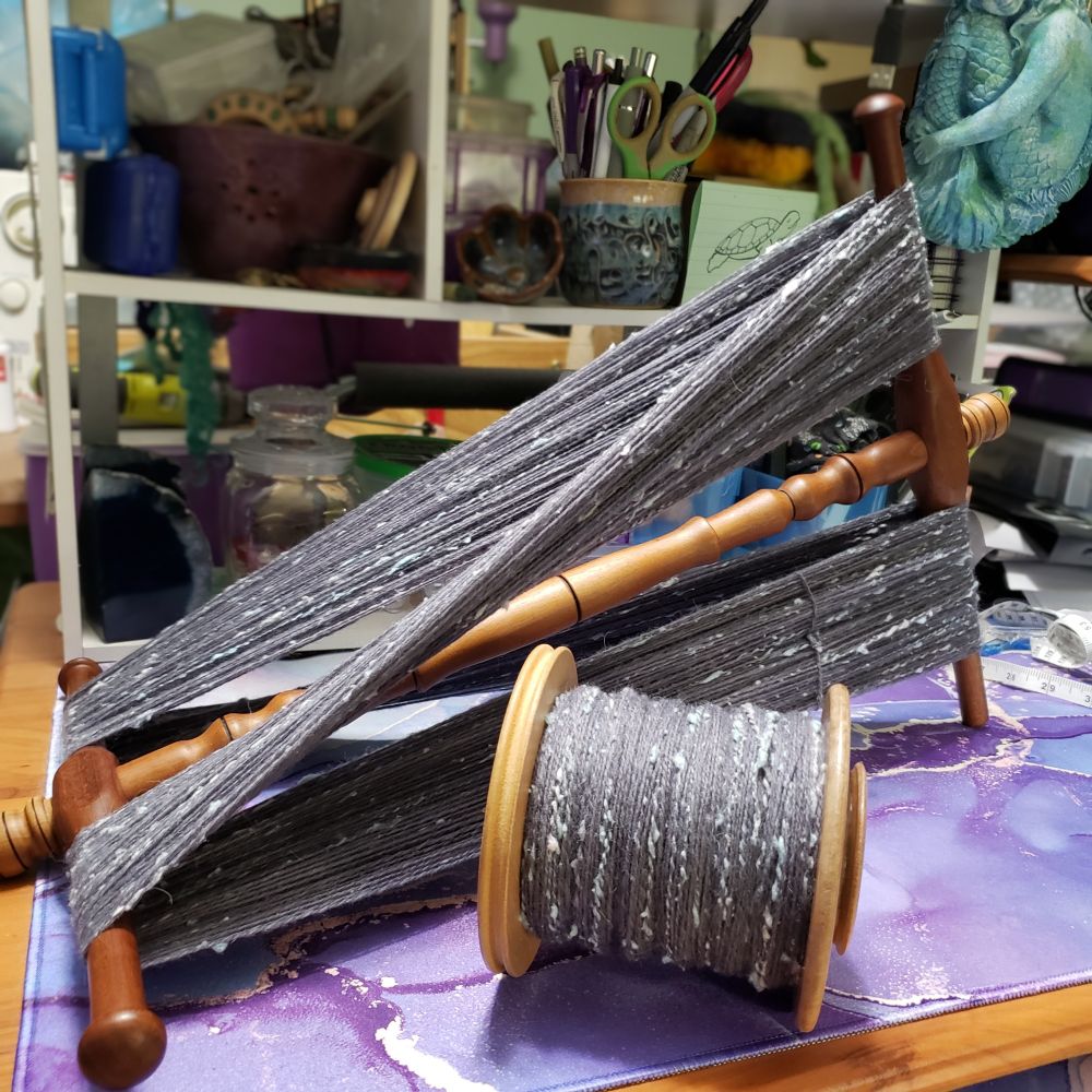 A view of Kelli's desk on which sits a hand turned wooden niddy noddy and an Ashford jumbo bobbin. Both are holding handspun two ply yarn that is primarily medium dark grey targhee wool and llama fiber along with pale silver grey and pastel blue silk noil. 