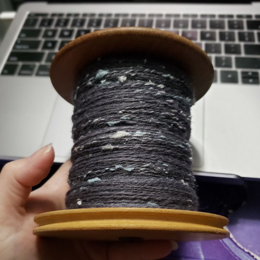 Kelli holds up an Ashford jumbo bobbin in front of their laptop. The bobbin is about half full of handspun two ply yarn that is primarily medium dark grey targhee wool and llama fiber with pale silver grey and pastel blue silk noil.