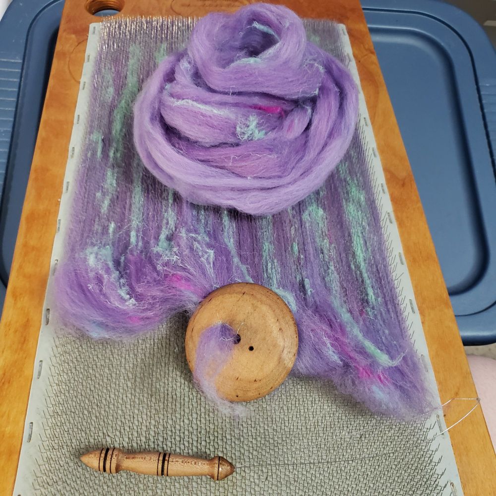 An overhead view of Kelli's blending board balanced on top of a storage tote. The spinning fiber that has been blended on the board is in the process of being pulled off through a diz into roving. A hand turned wooden diz and threader are on the foreground and a nest of roving that has been pulled off of the board rests in the background. The fiber is primarily targhee wool that has been immersion dyed in a range of lavenders with added hemp fiber in pale sea green, magenta and pale aqua blue. 