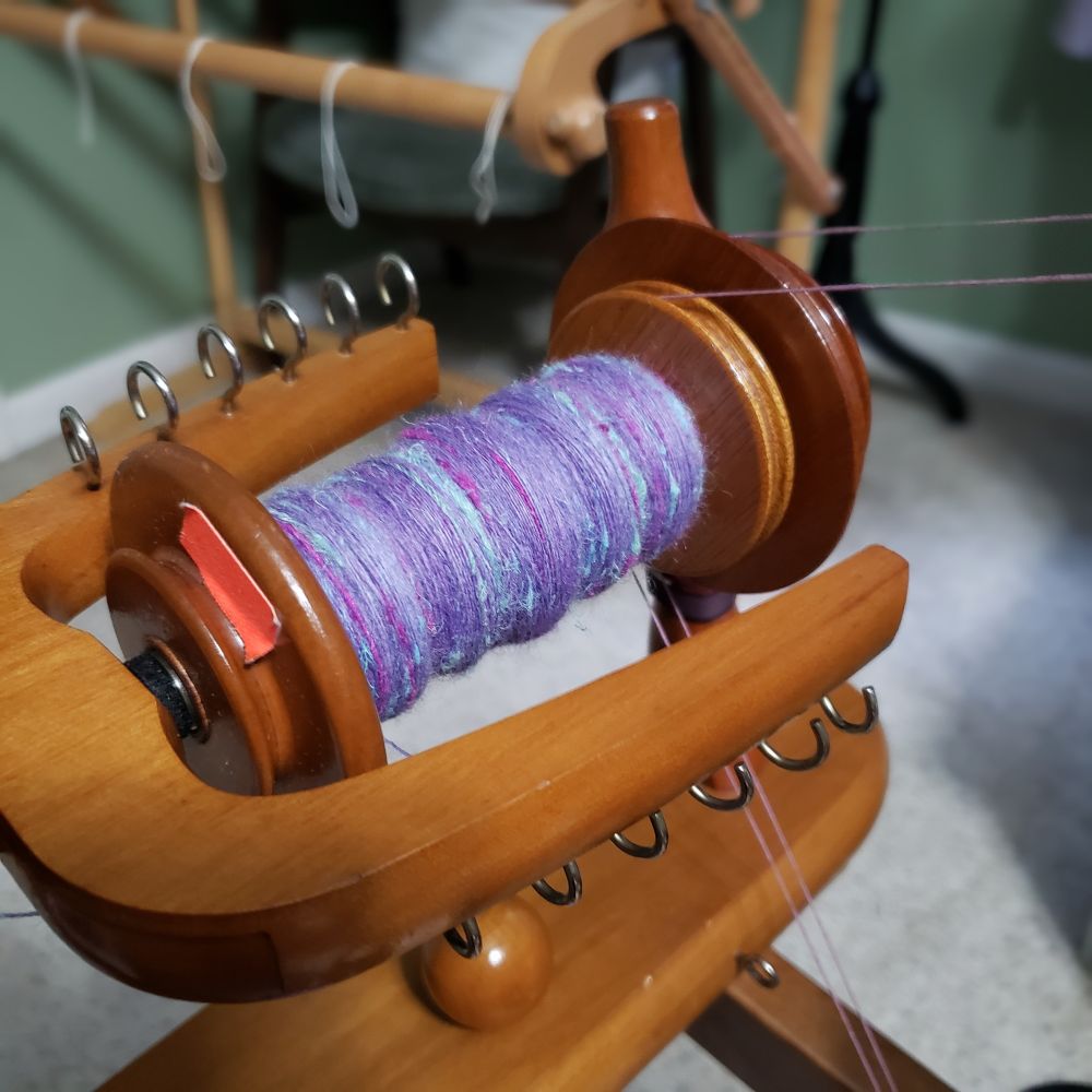 A closeup view of Kelli's Ashford Traditional spinning wheel. The bobbin is partially full of handspun singles that are primarily a range of lavenders with added hemp fiber in pale sea green, magenta and pale aqua blue. 