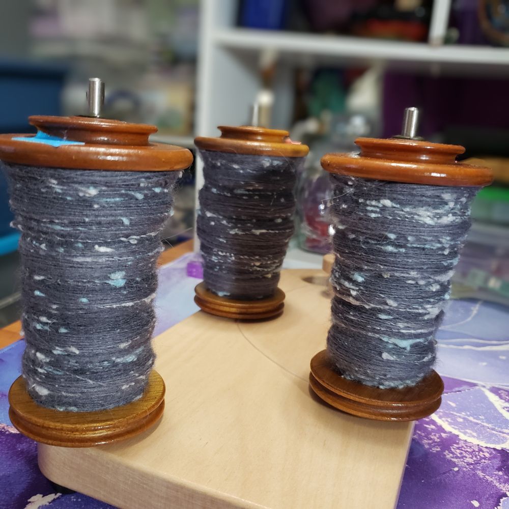 A closeup view of Kelli's Ashford tensioned lazy kate with three bobbins of handspun singles that are primarily medium dark grey targhee wool and llama fiber along with pale silver grey and pastel blue silk noil. 