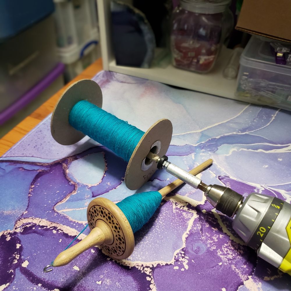 A hand turned wooden drop spindle with decorative pyrographed motifs on the top of the whorl sits on Kelli's desk. The spindle has a small cop of handspun turquoise singles yarn wound onto the shaft. A cardboard storage spindle with more of the same color handspun singles wound onto it is also laying on the desk attached to a wireless drill. The drill has just been used to wind the bobbin. 