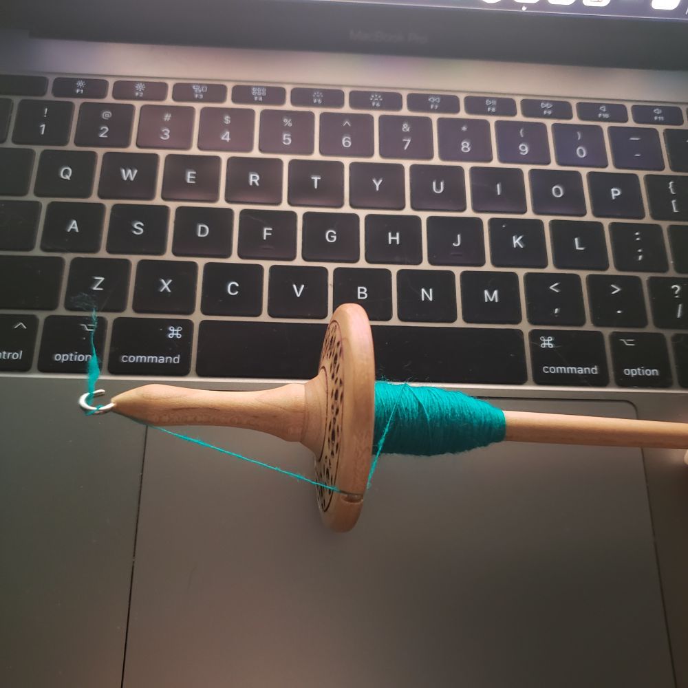 A hand turned wooden drop spindle with a small amount of turquoise wool handspun singles wound onto it sits across a laptop keyboard. 
