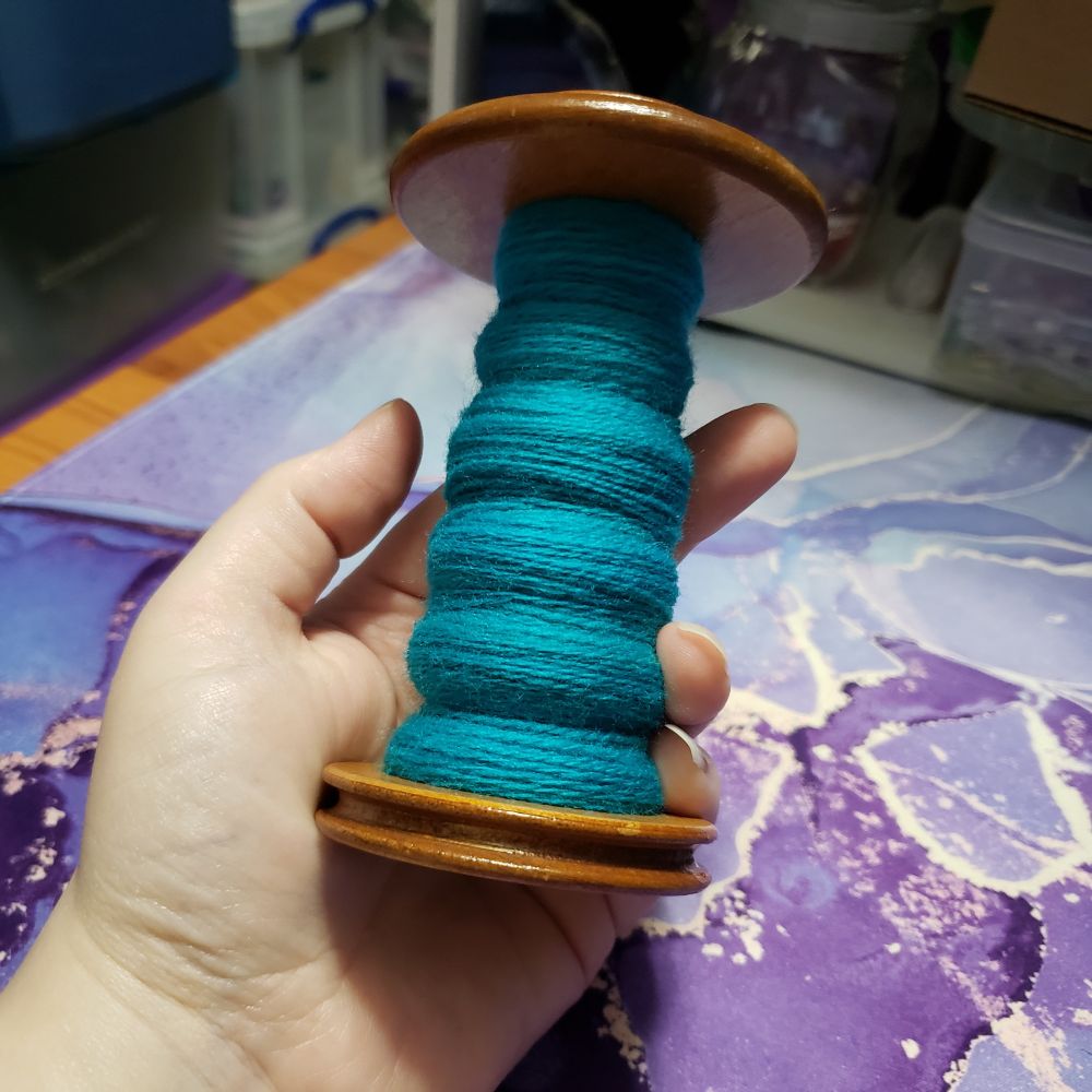 Kelli's hand holds up an Ashford standard spinning wheel bobbin in front of their desktop. The bobbin is partially full of turquoise two ply handspun superwash merino yarn. 