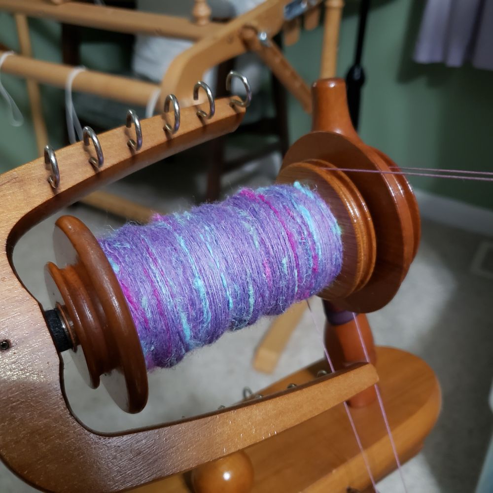 A closeup view of Kelli's Ashford Traditional spinning wheel. The bobbin is mostly full of handspun singles that are primarily a range of lavenders with added hemp fiber in pale sea green, magenta and pale aqua blue. 