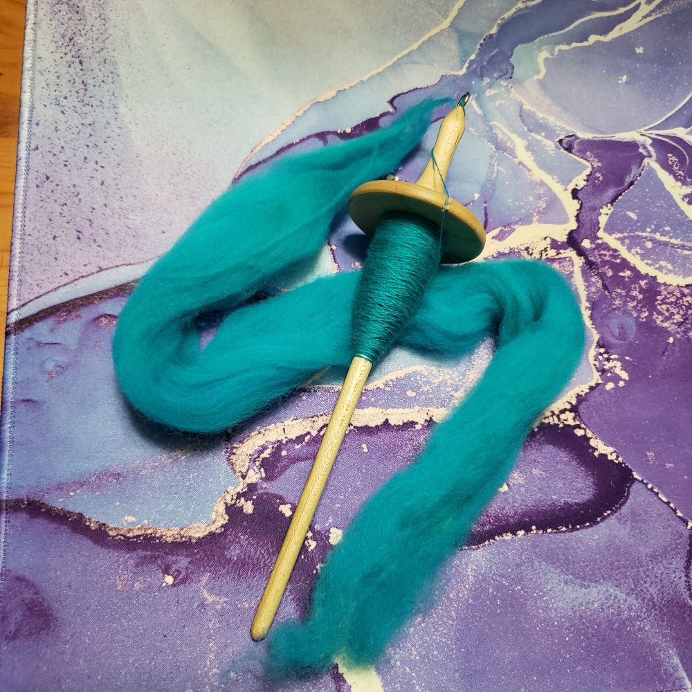 A hand turned wooden drop spindle sits on Kelli's desk. It is partially full of handspun singles yarn that has been hand dyed turquoise. Under the spindle is a small amount of turquoise superwash merino wool combed top which is in the process of being spun.