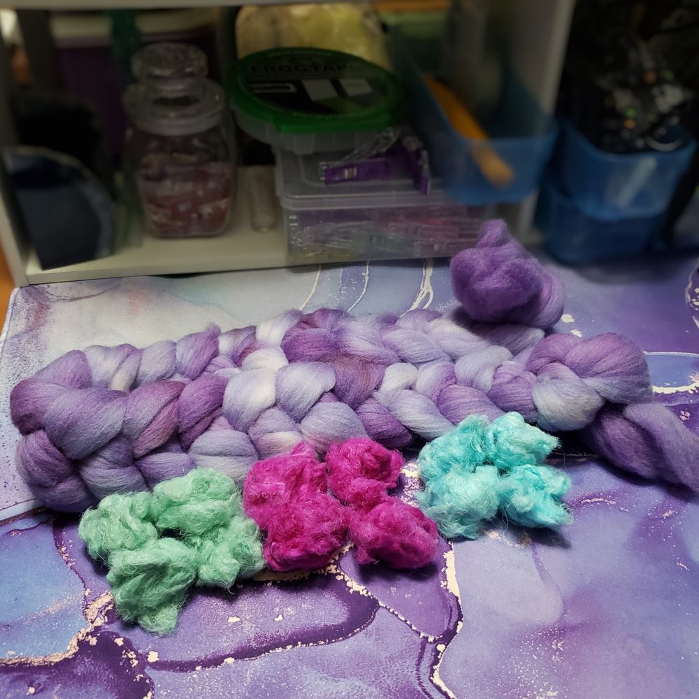 An immersion dyed braid of targhee wool in various shades of lavender sites on Kelli's desk. Tge desk mat is nearly the same combination of colors as the targhee. Three small piles of loose short hemp spinning fiber are sitting in front of the targhee. Each pile has been low water immersion dyed a different color. The left pile is a pale sea green. The middle pile is a medium fuchsia. The right pile is a pale aqua blue. 