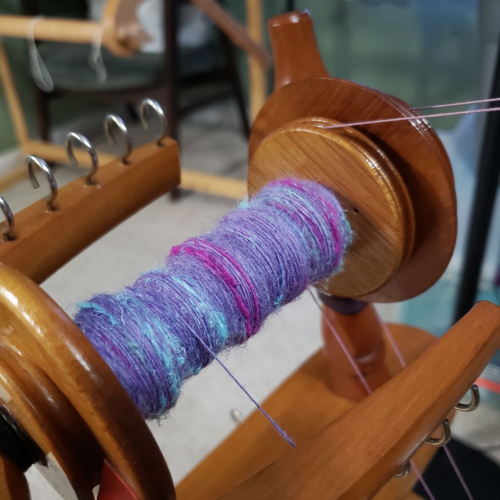 A closeup view of Kelli's Ashford Traditional spinning wheel. The bobbin is partially full of handspun singles which are primarily targhee wool that has been immersion dyed in a range of lavenders with added hemp fiber in pale sea green, magenta and pale aqua blue. 
