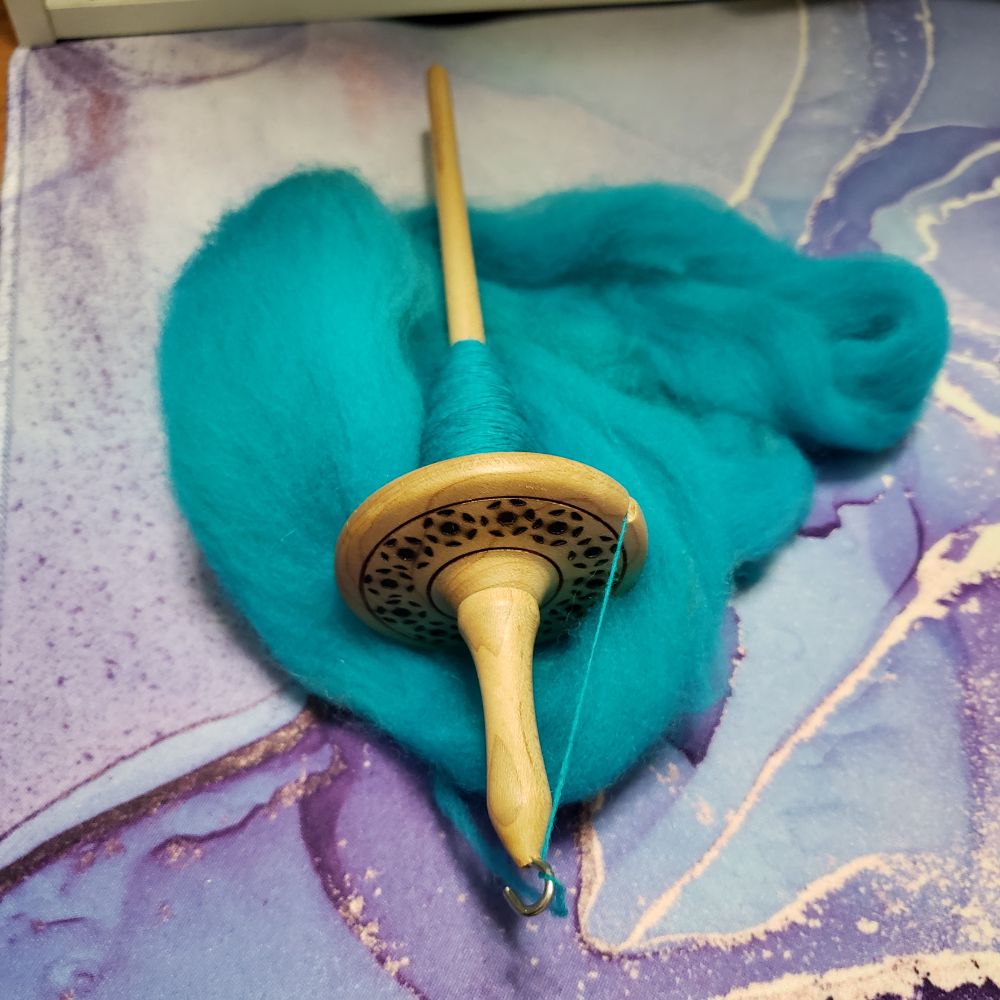 A hand turned wooden drop spindle sits on Kelli's desk. It is partially full of handspun singles yarn that has been hand dyed turquoise. Under the spindle is a small amount of turquoise superwash merino wool combed top which is in the process of being spun. The top of the spindle whorl has decorative pyrographed motifs. 