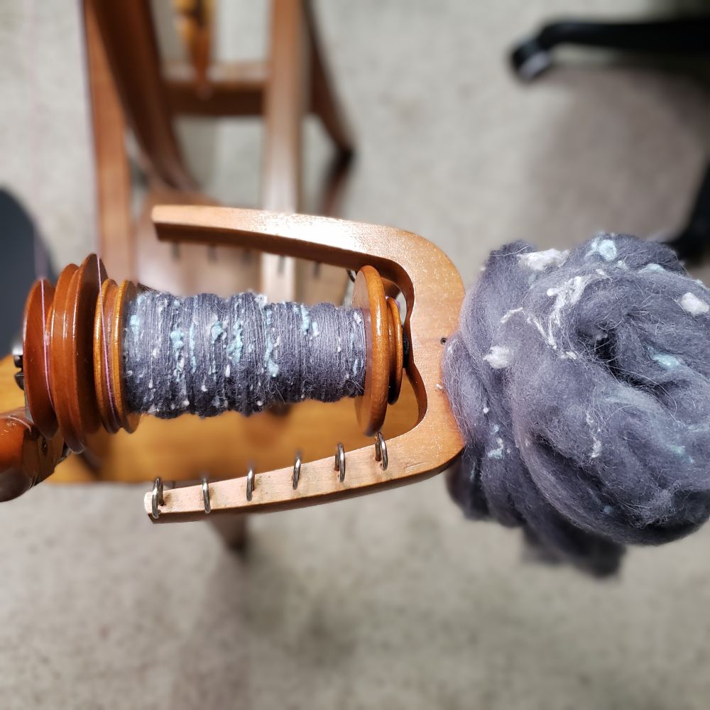 An overhead view of Kelli's Ashford Traditional spinning wheel. The bobbin is partially full of handspun singles that are primarily medium dark grey targhee wool and llama fiber along with pale silver grey and pastel blue silk noil. A nest of the blended spinning fiber sits on the front maiden upright. 