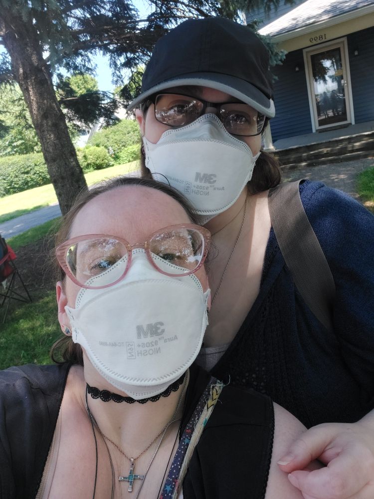 Kelli stands behind their Wife, Rae. Both smile behind their N95 masks and are watching the Rochester Pride Parade. A random house along the parade route is in the background. 