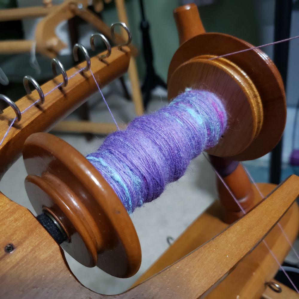 A closeup view of Kelli's Ashford Traditional spinning wheel. The bobbin is partially full of handspun singles that are primarily targhee wool that has been immersion dyed in a range of lavenders with added hemp fiber in pale sea green, magenta and pale aqua blue. 