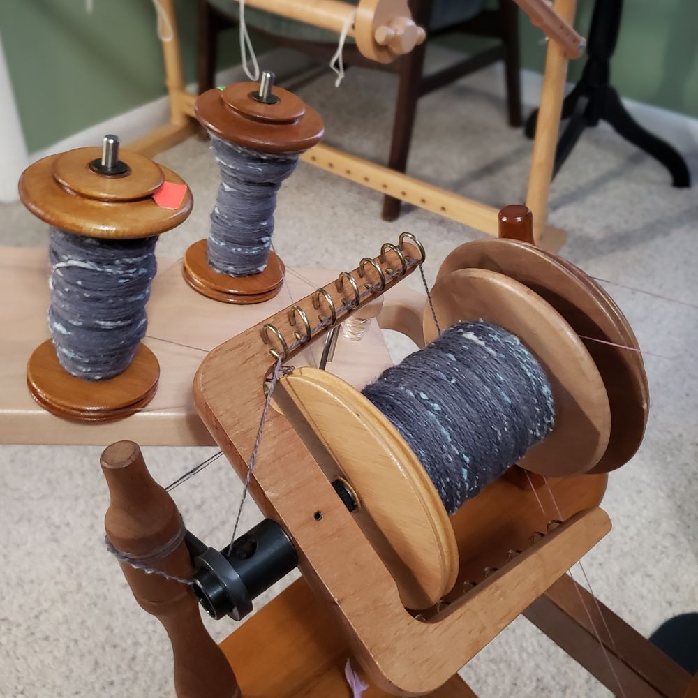 An angled closeup of Kelli's Ashford Traditional spinning wheel equipped with a jumbo flyer. Kelli holds up an Ashford tensioned lazy kate behind the wheel's flyer to show off two partial bobbins of handspun singles. The jumbo bobbin is partially full of the resulting two ply handspun yarn. The yarn is primarily medium dark grey targhee wool and llama fiber along with pale silver grey and pastel blue silk noil. 