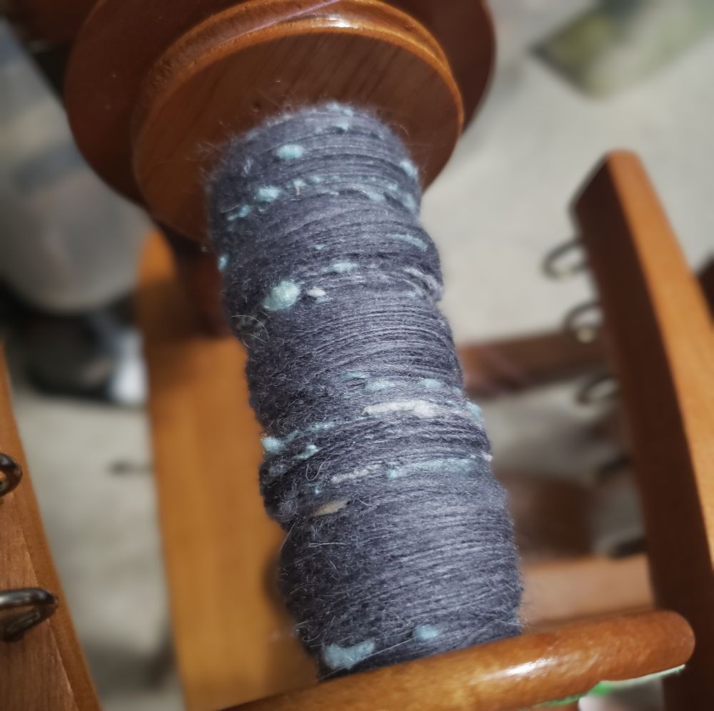 An overhead view of Kelli's Ashford Traditional spinning wheel. The bobbin is partially full of handspun singles that are primarily medium dark grey targhee wool and llama fiber along with pale silver grey and pastel blue silk noil. 