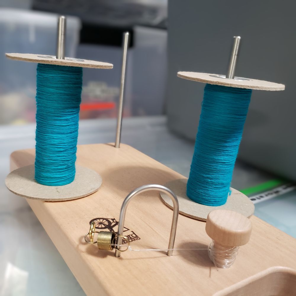 An Ashford tensioned lazy kate holds two cardboard storage bobbins that each have a small amount of turquoise handspun singles yarn on them. 