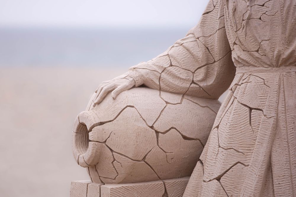 A closeup of a creation or statue made of sand. In this creation a vase lies on its side with a woman's hand placed on it. The empty vase and left side of the woman are cracked and broken. Behind the statue is a sandy beach and distant Atlantic Ocean during low tide at Hampton Beach, New Hampshire, USA. This piece is entitled "It Won't Stay Forever" by artist Dmitry Klimenko constructed at the 25th Hampton Beach Sandcastle Competition  