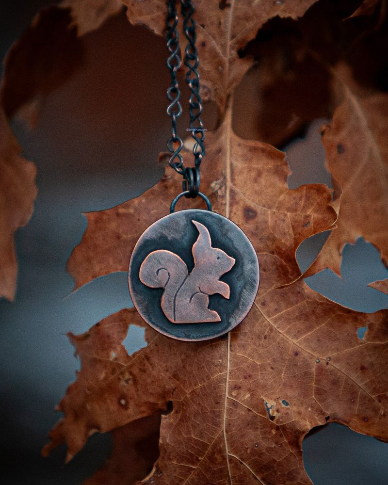 a round necklace on a chain with the side view of a squirrel formed from copper that has been darkened with an artistic patina. It's resting on a dried red oak leaf, and there is snow on the ground in the background creating a white mottled look under the tree.