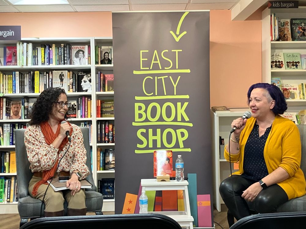 Venessa Vida Kelly and Alex Villasante talking about FIREBLOOMS in front of a black and green sign that says East City Books. 