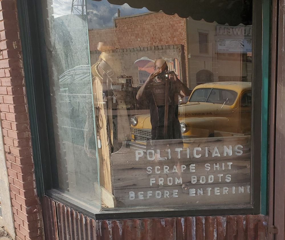 Me, with a1940s yellow Chrysler, reflected in a window that reads: politicians scrape shit from boots before entering