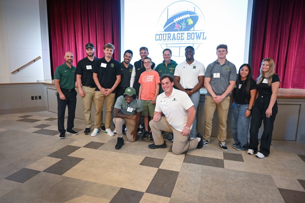 Members of SUNY Brockport's football team and cheerleading squad pose for a photo in Cleary Family Auditorium with an honorary coach, in front of the Courage Bowl logo.