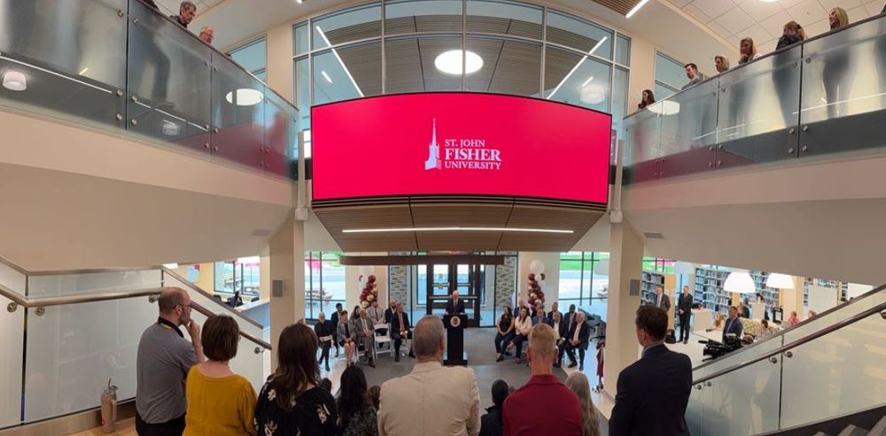 Underneath a screen featuring the St. John Fisher University logo, members of the Fisher community sit on the Social Stairs of the renovated and modernized Lavery Library to celebrate its grand reopening