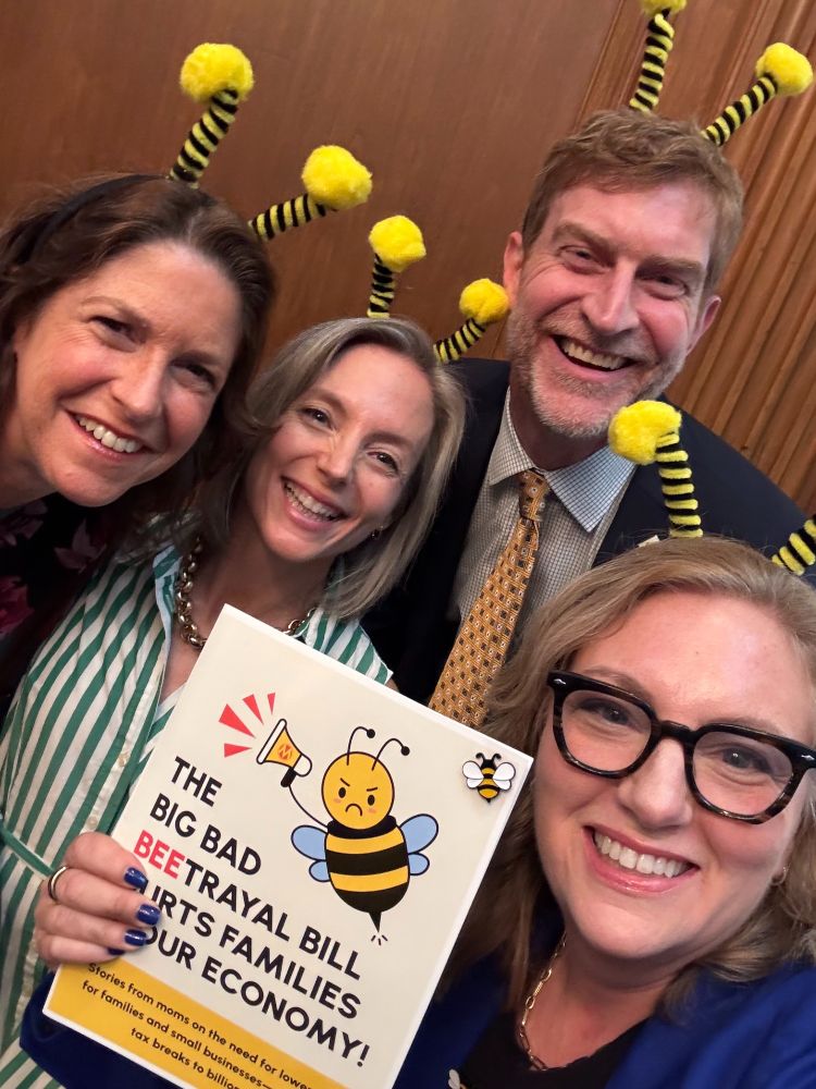 Four people wearing bee antennae headbands smiling at the camera and holding a book. The title of the book is ""The big bad beetrayal bill hurts families and our economy" and has a drawing of a bee holding a megaphone and looking angry. 
