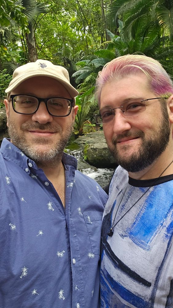 This image features two men smiling at the camera in what appears to be a lush, tropical setting with greenery and rocks visible in the background, suggesting a natural environment like a rainforest or botanical garden.