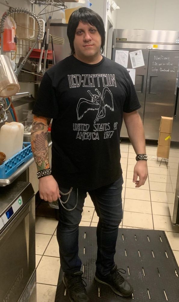 A man with a black wig, wearing a black Led Zeppelin "United States of America 1977" t-shirt, dark jeans, and black sneakers stands in what appears to be a commercial kitchen. He has a tattooed left arm and is wearing spiked bracelets on both wrists and a chain connected to his jeans.